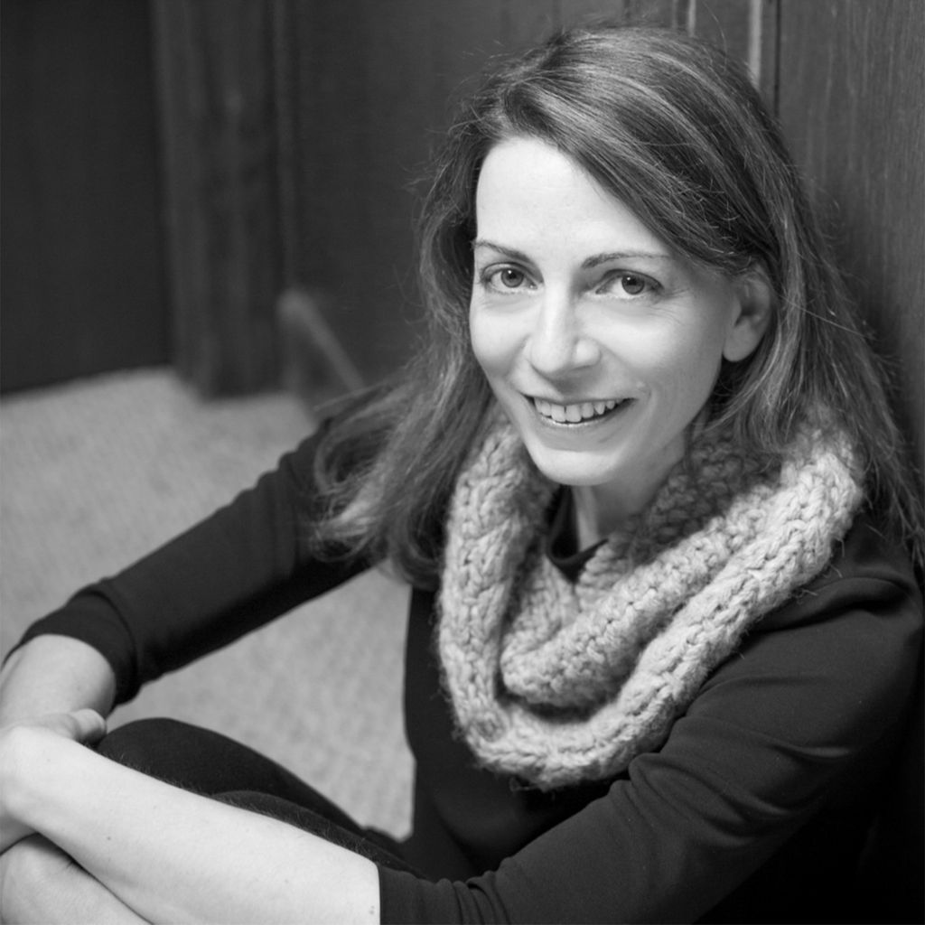 A black and white portrait of Susan Barba (GRS’12) in a scarf, smiling as she sits on the ground.