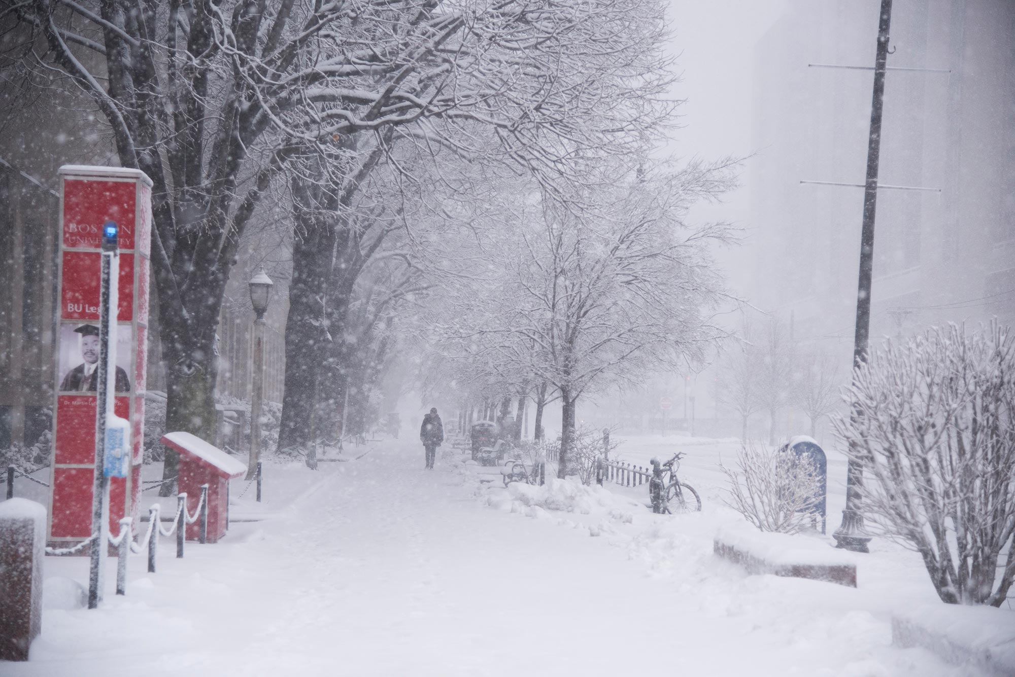 Photo taken during a Nor'Easter in 2018; the sidewalk is covered with snow as figure in jacket walks down. Snow falling blurs the image.