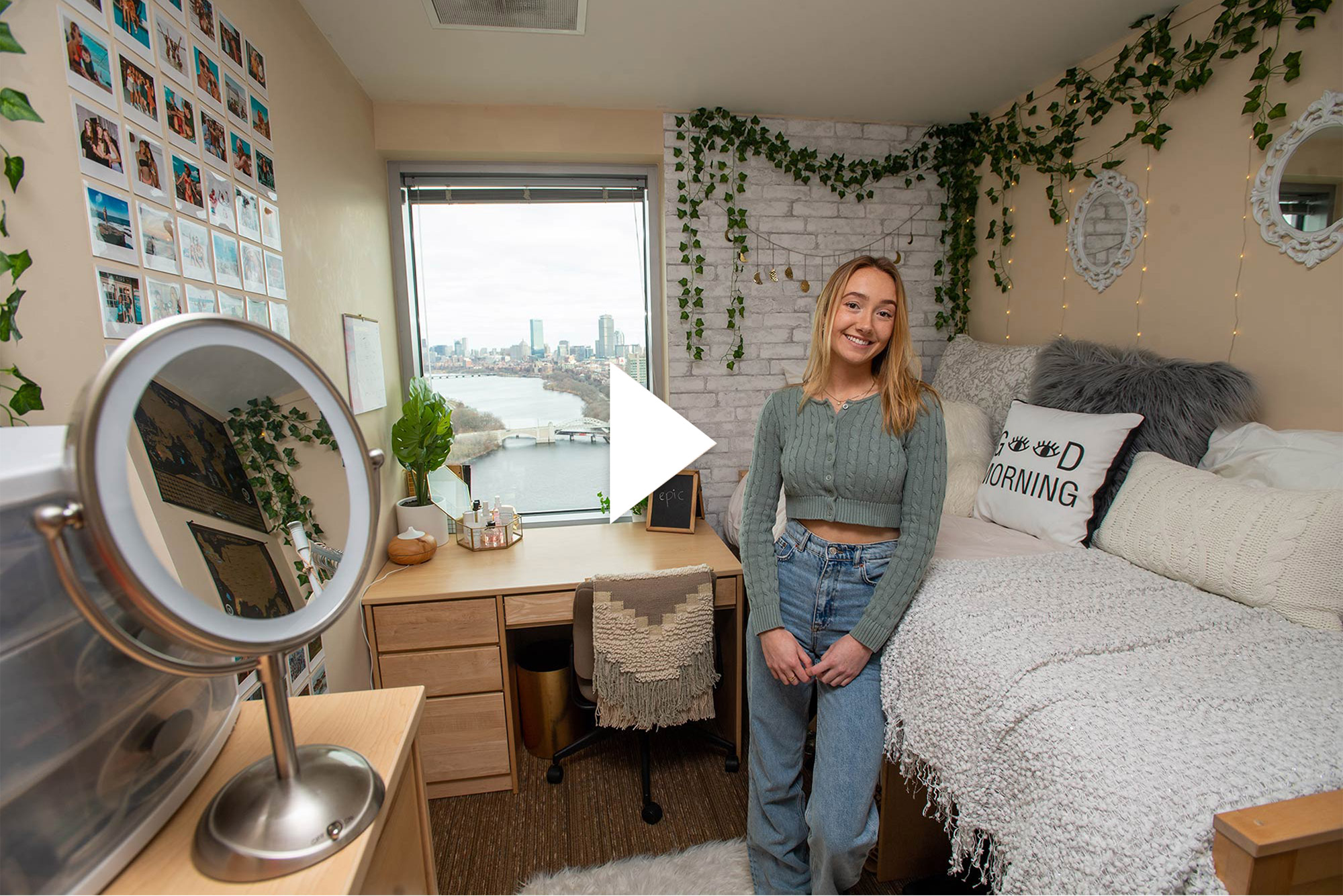 Photo of Casey Evans (COM’21) standing in her StuViII quad with a million-dollar view of Boston’s skyline in the window at the back of the room. The dorm room is beautifully decorated with hanging plants, a wall of photos, and a bed with many pillows, one of which reads “good morning.”