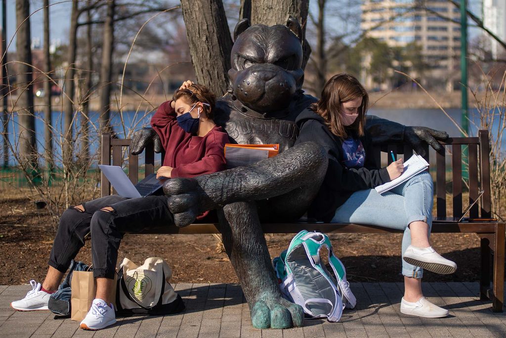 Photo of Alice Hotimski (CGS’22,CAS’24), left, and Eleanor Paul (CAS’24) get some work done in some beautiful 70 degree weather on March 11 as they sit on either side of the Rhett bench. They wear face masks and sit with their laptops and notebooks in their laps.