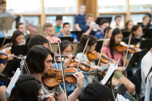 Photo of a small orchestra with student musicians rehearsing. In the bottom left corner, William Peltz-Smalley (CFA'22), who has brown, ear-length hair, is seen in focus with his bow balanced on his violin’s strings. In the background, other students with string instruments are blurred.