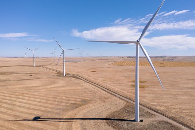 Photo of large white wind turbines in a dry, flat landscape. A blue sky with small clouds are seen.