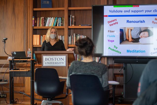 Photo of Paula Quatromoni (SPH’01) standing at a Boston University Podium at the front of classrom with a slide show behind her that shows a picture of a person grabbing their stomach, with the words “Validate and support your client” above it and the words “dietary variety, renourish, fiber, fluids, yoga, try probiotics, meditation” around it. Quatromoni wears a patterned face mask and a few blurred students are seen sitting in the classroom.