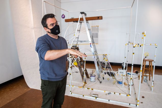 Photo of CFA’s David Snyder, in a blue long sleeve and black face mask, at the 2021 MFA Thesis Sculpture Exhibit. He stands in front of an untitled piece by  Aris Hu (MFA’21), which consists of a structure made of pipes that contains different sizes of ladders and stools.