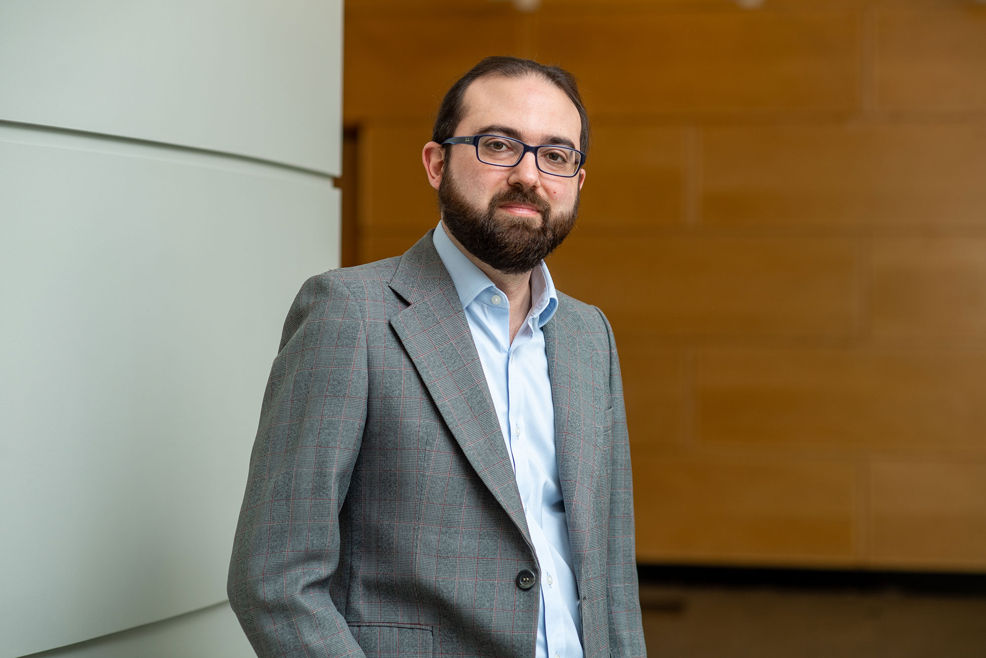 Photo of Gianluca Stringhini (ENG) looking serious, with his hand in his pocket, wearing a gray blazer and standing in a hallway with a wooden wall in the background.