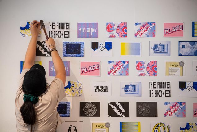 Photo of Jenna Benoit (CFA’21) helping install part of this year’s MFA Graphic Design Thesis Exhibition, The Printer Is Broken, on view through April 6 at CFA’s Faye G., Jo, and James Stone Gallery. She is seen with a hammer from behind, working to hang up a piece of paper that says "the printer is broken" amongst other colorful papers.