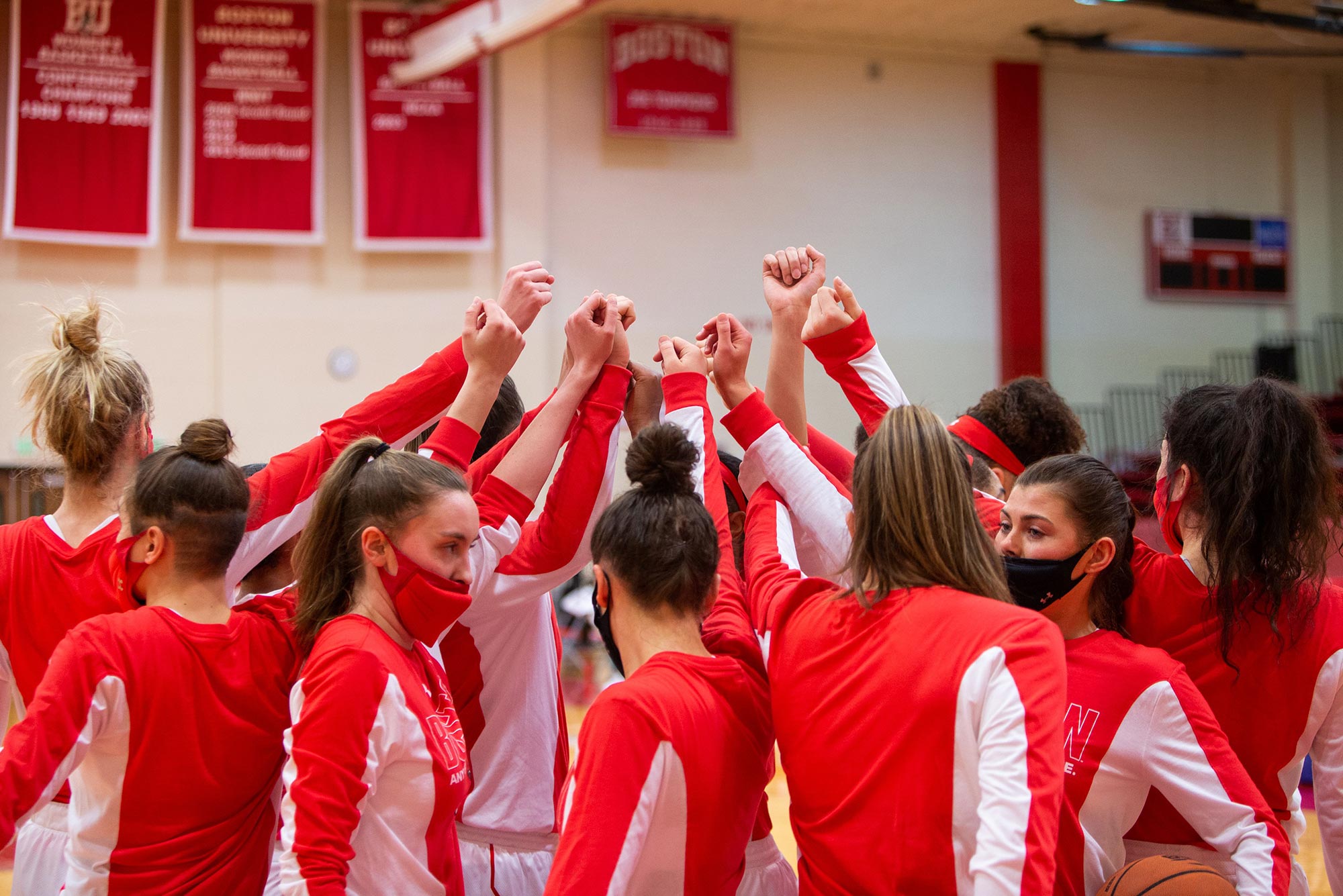 Photo of the BU women’s basketball team with their hands raised in the air during what looks like a time out. They wear red and white long sleeve shirts and face masks.