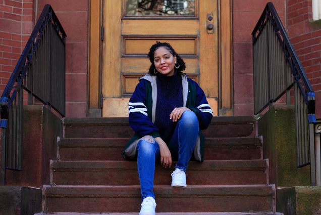 Photo of Solange Hackshaw (COM’21) in a stylish blue coat with white stripes on the sleeves sitting on the steps outside of a brown stone. We smiles, wears hoop earrings and rests her hands on her knees.