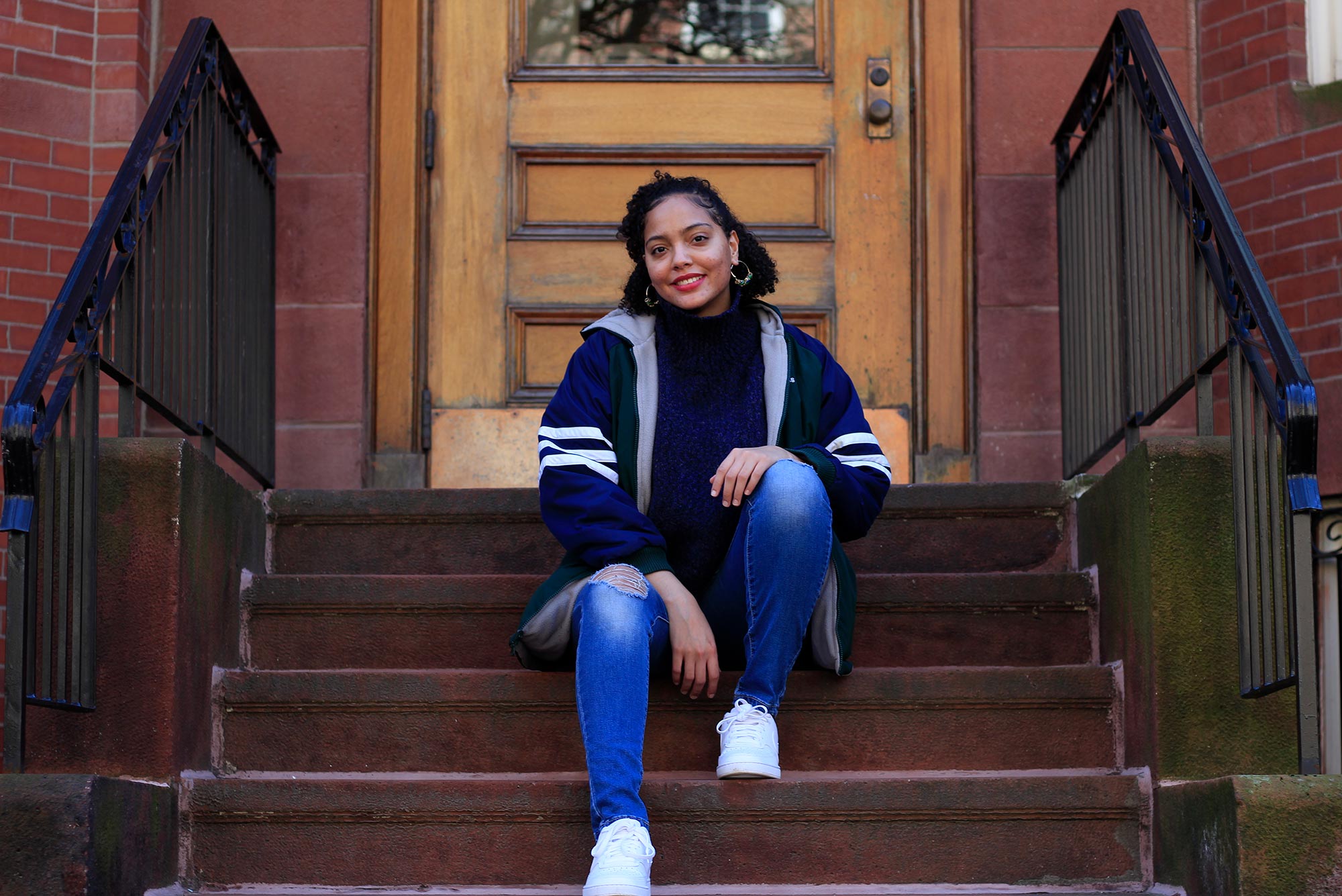 Photo of Solange Hackshaw (COM’21) in a stylish blue coat with white stripes on the sleeves sitting on the steps outside of a brown stone. We smiles, wears hoop earrings and rests her hands on her knees.