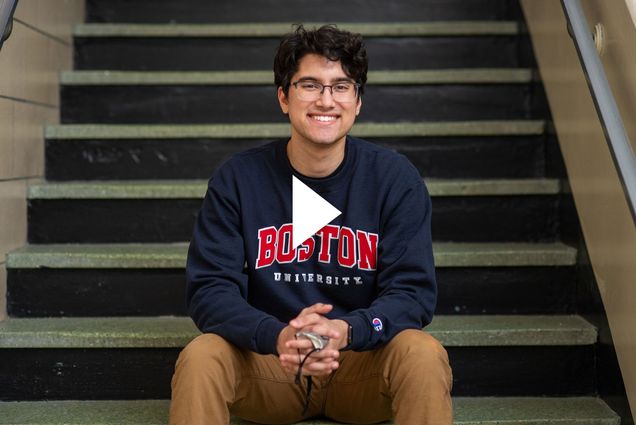 Photo of student vlogger Amit Bhatia (Questrom'24) sitting on the steps of a staircase with a navy blue sweatshirt that says “Boston University” in red and white. He smiles and holds his face mask in his hands. A white video play button is overlaid.