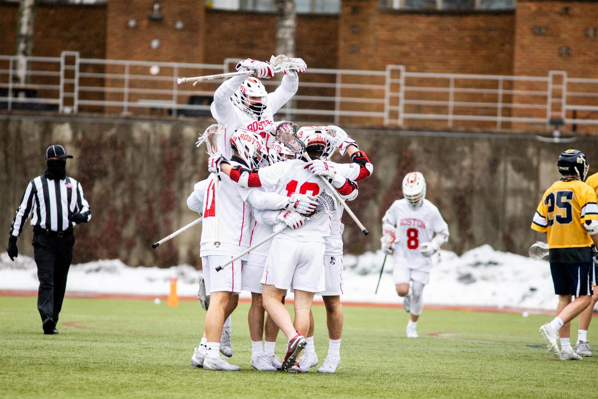 Photo of the a huddle of Men’s lacrosse players, dressed in white, in a celebratory huddle, during their game against Pace University on February 27 at Nickerson Field. A referee and members of the Pace team are seen in the background.