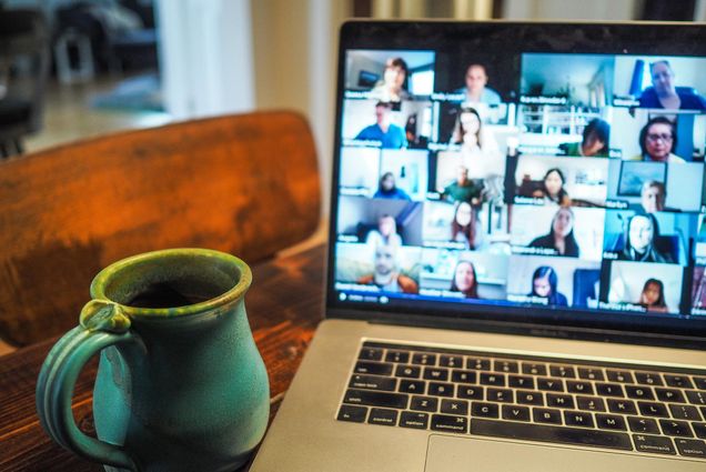 Photo of a teal mug resting on a wooden table next to MacBook with a blurred video call with more than a dozen participants on screen.