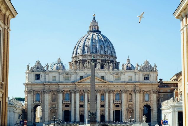 Photo of St. Peter’s Basilica in Vatican City on a sunny day; a seagull flies by in the top right corner.