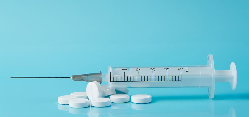 Photo of a pile of white, circular pills with a syringe resting on top; the background is completely light blue. Semaglutide can be administered in both forms.