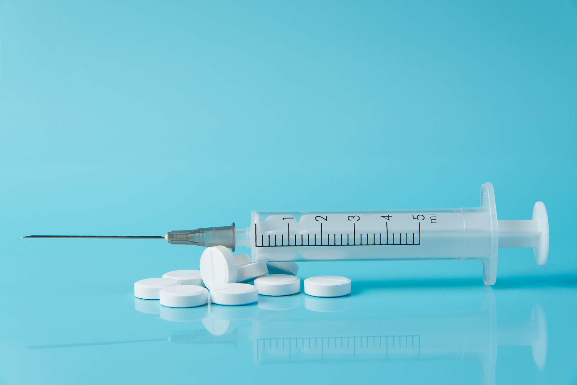 Photo of a pile of white, circular pills with a syringe resting on top; the background is completely light blue. Semaglutide can be administered in both forms.