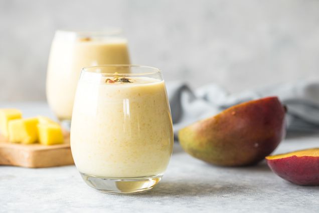 Photo of a short tumbler glass filled with a yellow colored Mango Lassi. In the background, a cutting board with mango pieces and a mango cut in half sit on the counter.
