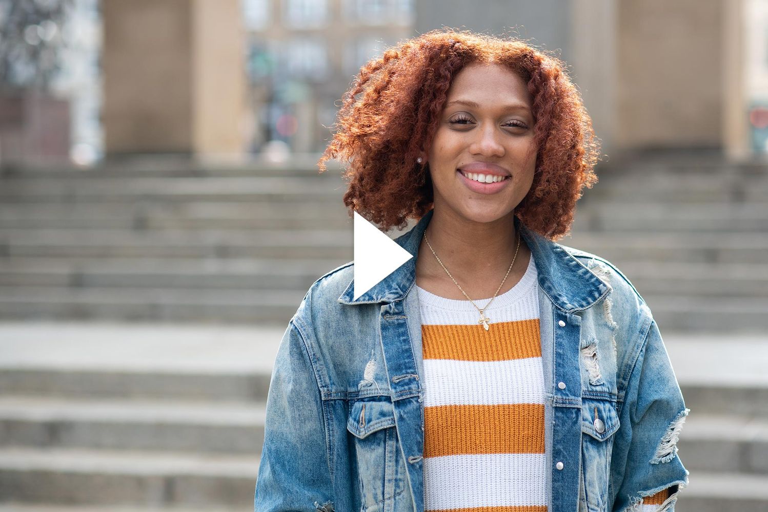 Photo of Nyah Jordan (CGS’20, COM’22) standing in front of the steps in Marsh Plaza. She smiles, has curly shoulder-length reddish hair, and wears a jean packet and gold and white striped sweater.