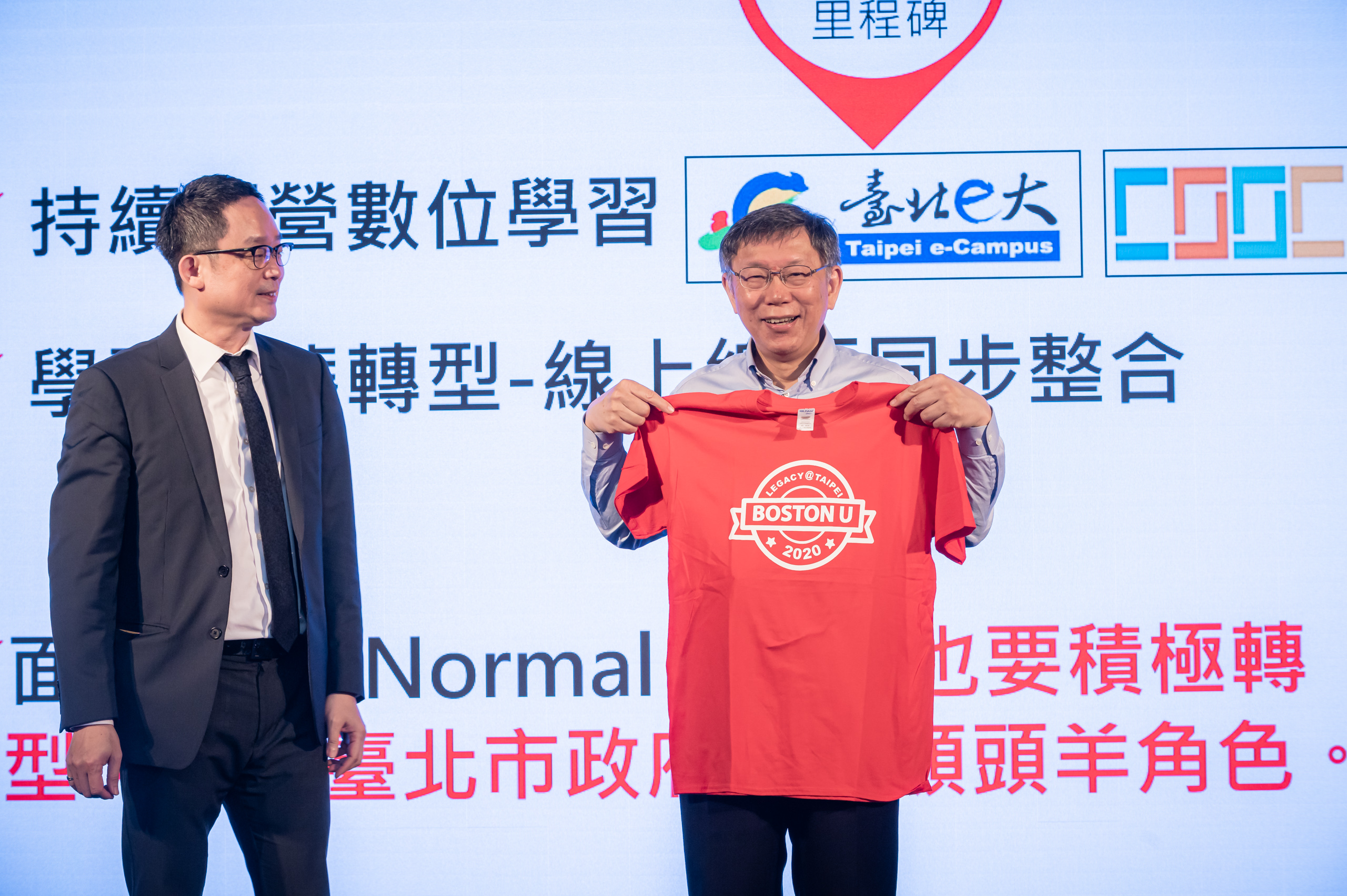 Photo of Vincent Hsieh (LAW’92) (left) and Ko Wen-Je, the mayor of Taipei (right), who holds up a red t-shirt that says “Boston U” on it. Behind him, a large screen with Chinese characters is seen.