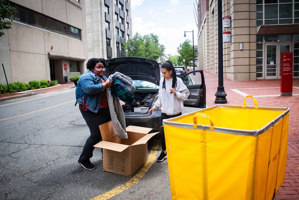 Photo of Robyn Volcy-Lee (left) and Katerina Chew as they pack up their car on June 1, 2020. A yellow cart is seen in the right foreground, Lee removes some clothes from a cardboard box on the ground.