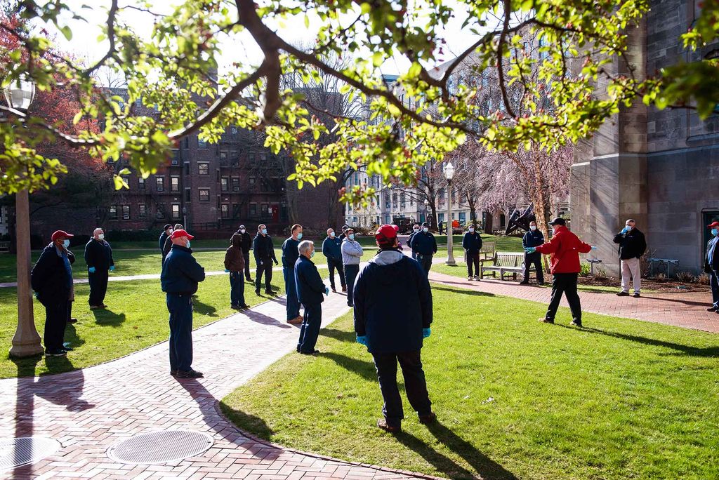 Elijah Ercolino, director of Building Automation Systems for Facilities Management & Planning, dressed in red at right, holds a meeting outside of the School of Law on April 29, 2020. The team are all spaced out 6 feet apart, many wear red baseball caps. The branch of a spring tree is seen in the foreground at top.