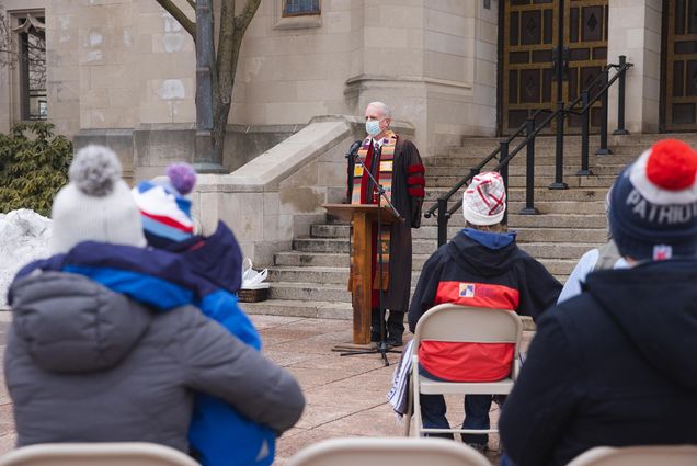 Photo of Rev. Dr. Robert Allan Hill, Dean of Marsh Chapel, leads a morning prayer service Sunday morning at Marsh Plaza. He stands at the front of a group in winter coats and hats sitting in folding chairs, and wears a mask as he speak into the podium microphone.