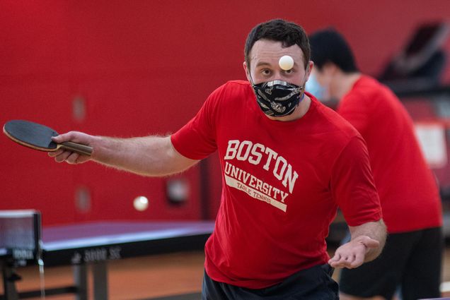 Photo of Ryan Hong (QST’23) in a red Boston University shirt mid swing during a game of table tennis with his club team at FitRec January 21. He intently watches the ball and wears a black and white face mask.