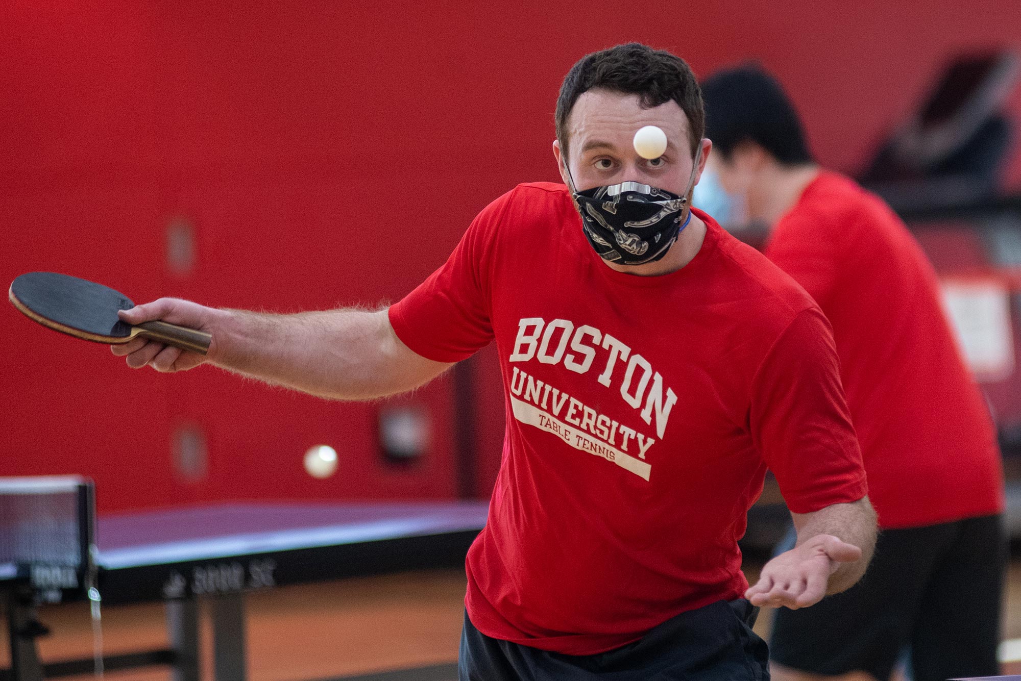 Photo of Ryan Hong (QST’23) in a red Boston University shirt mid swing during a game of table tennis with his club team at FitRec January 21. He intently watches the ball and wears a black and white face mask.