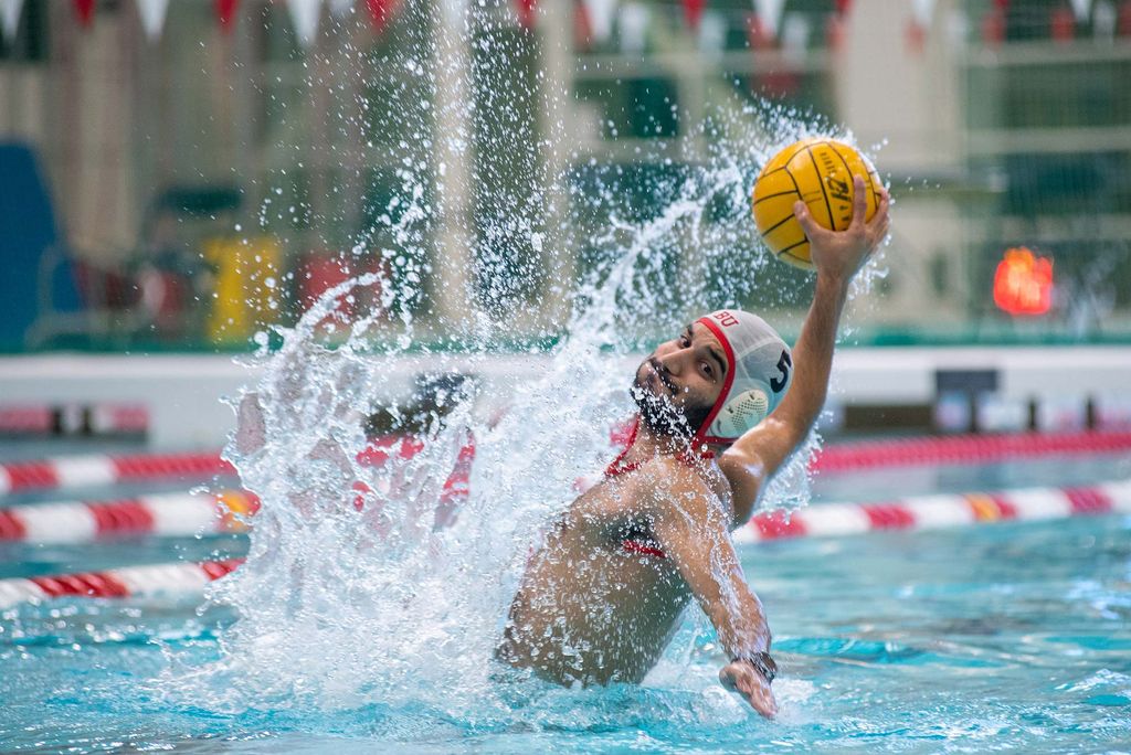 Photo of Laith Hijazi (QST’23) with his lips pursed as he stretched back to throw a water polo ball to one of his teammates on his club team at FitRec January 21. A huge splash of water is seen to his right and behind him, and the red and white floating lane dividers of the pool are seen in the background.