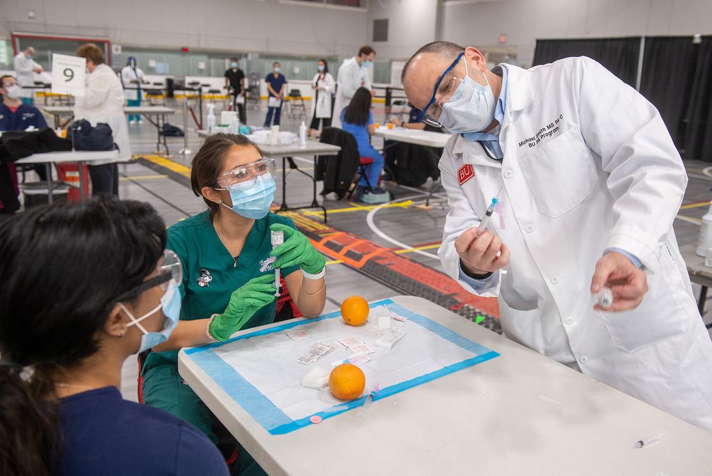 Photo of BU med student Alekha Kolli, from left, and Tia Patterson (DMD’24) learning from Michael Smith, MS PA-C at the vaccination administering training at FitRec March 7. They all wear face masks and goggles and sit around a table in the middle of the gym.