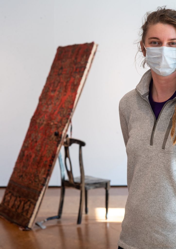 Photo of Hannah Minife (MFA’21) standing in front of  part of her sculpture,  “Three Doors.” She wears a gray pullover and blue face mask. Her sculpture features a door covered with an elaborate textile pattern, balancing against a chair, with an axe resting on the ground. 