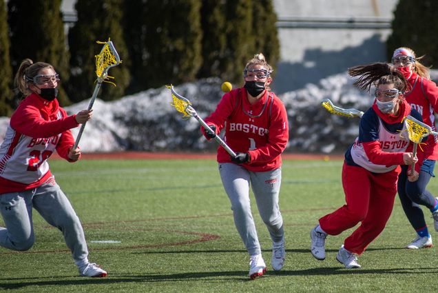 Photo of a women’s lacrosse team practice on Nickerson Field March 5, 2021. Makenzie Irvine (CAS’21), from left, Quinn Ferry (CAS’24), and Ava Barry (CGS’21) are seen in red Boston University sweatshirts and pinies; the yellow lacrosse ball is mid flight as two of the women reach for it with their sticks.
