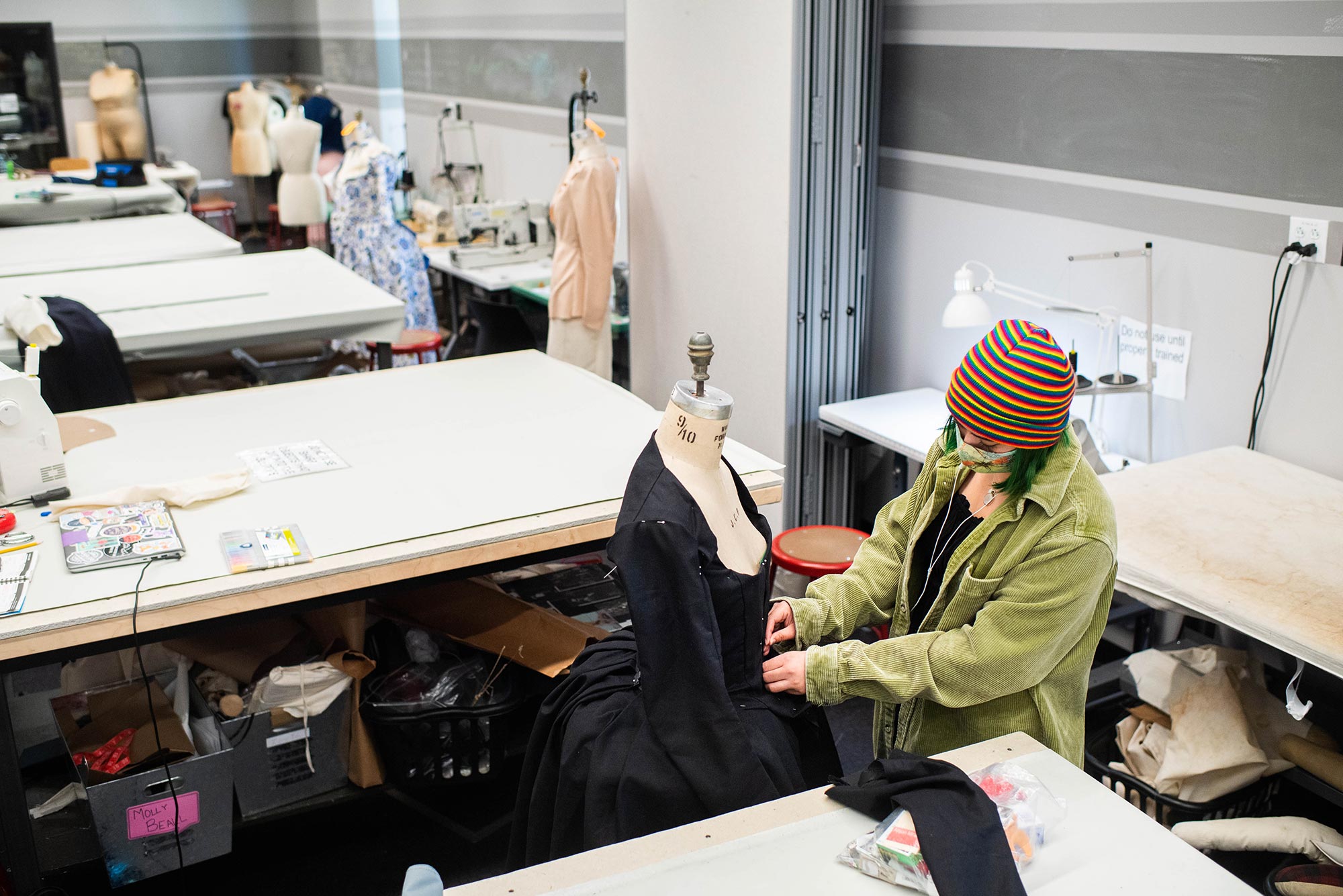 Photo of Alison Miranda (CFA 22) working on pining a black dress with a long skirt during a costume production class at the Booth Theater on March 15, 2021. Miranda wears a colorful, rainbow striped hat and a face mask. Mannequins and work tables are seen in the background.