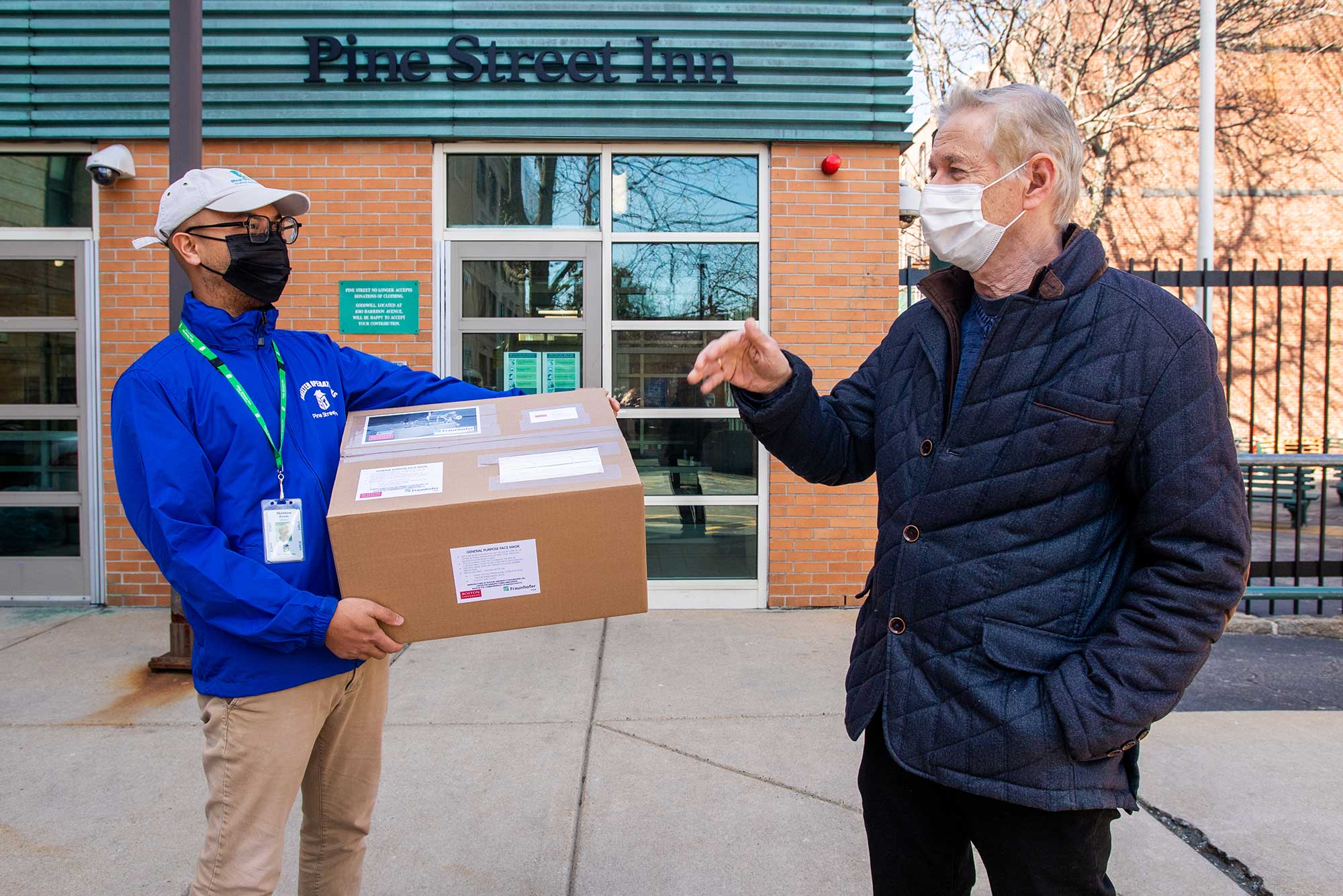 Photo of Andre Sharon (right), director of the Fraunhofer USA Center for Manufacturing Innovation at BU chats with Matt Ferrer, volunteer coordinator at The Pine Street Inn, after handing over a box of 1000 masks March 19. Behind them, a sandstone brick building with a dark blue “Pine Street Inn” sign is seen. Both men wear masks and Sharon raises his hand as he appears to explain something to Ferrer.