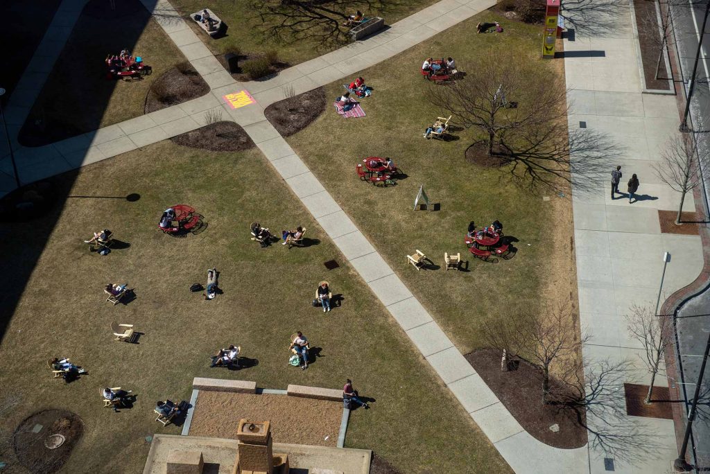 Aerial photo of the COM Lawn at a slight angle. The intersection of two cement paths is seen in the top left of the image, and the rest of the space is green and filled with people sitting on Adirondack chairs. A few people walk down the sidewalk on the right.