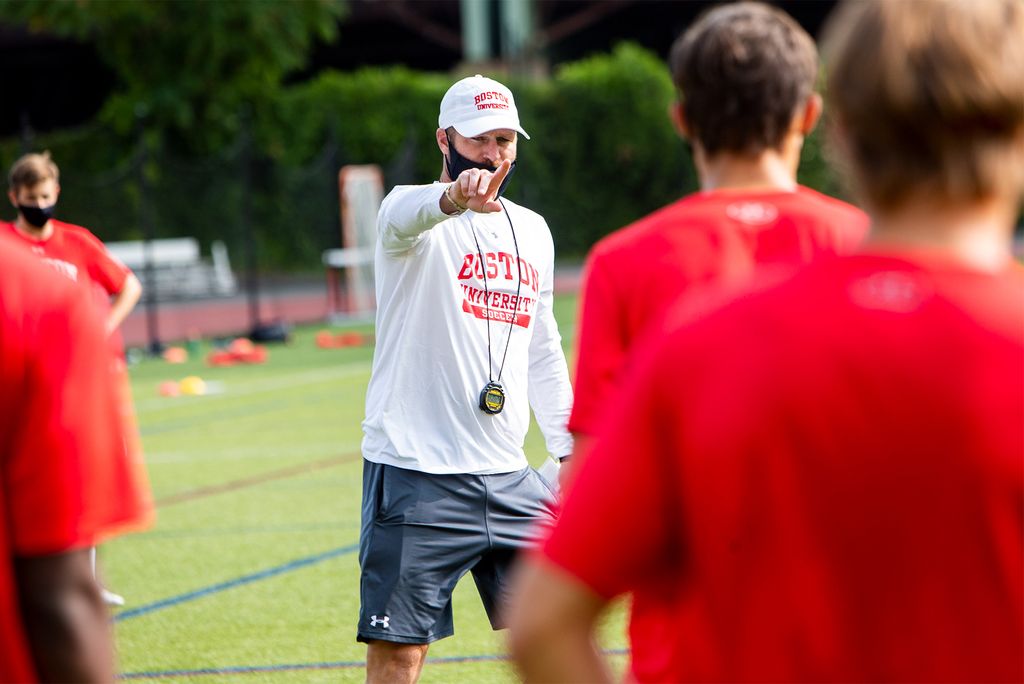 Photo of Kevin Nylen in a black face mask and white BU Athletics baseball cap and long sleeve pointing to members of the BU men’s soccer who are seen from the back, blurred in the foreground in red shirts.