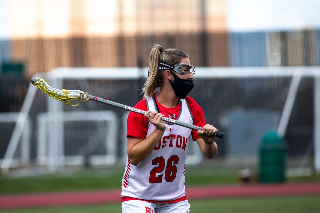 Photo of Makenzie Irvine in a red and white BU jersey with the number 26, reaches back with her lacrosse stick as she possess the ball. She wears eye protection and a black face mask.