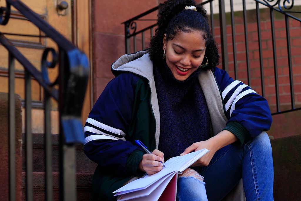 Photo of Solange Hackshaw (COM’21) in a stylish blue coat with white stripes on the sleeves sitting on the steps outside of a brown stone, smiling as she writes in a notebook.