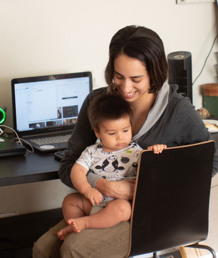Photo of a young person smiling, with a baby in their lap, turned away from their computer as they sit at their desk.