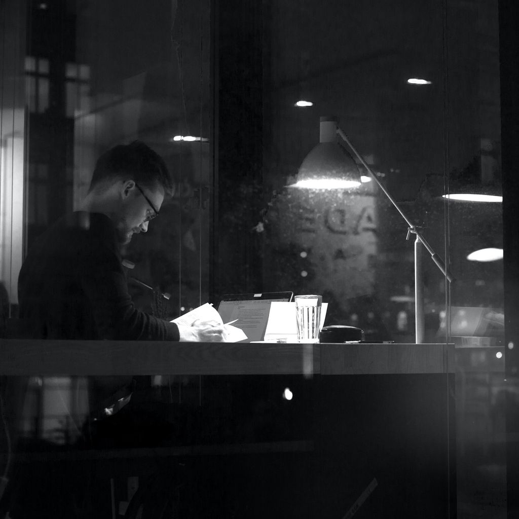 Black and white photo of a young man working at a desk with a book in his hand with a bright desk lamp shining down on his book.
