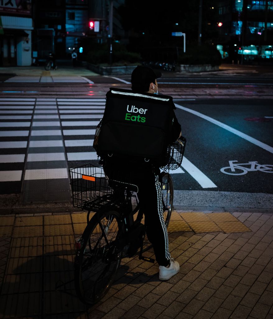 Photo taken from behind a person on a bike with a rear bike basket who waits at a crosswalk. They have an “Uber Eats” insulated backpack on.