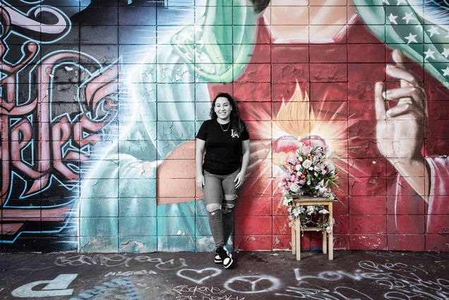 Photo of Daisy Figueroa (LAW’21) in a black t-shirt and jeans ripped at the knee standing if front of a larger mural with the virgin mary and graffiti painted all over it. The photo has been edited so Figueroa is in black and white and the mural is brightly colored.