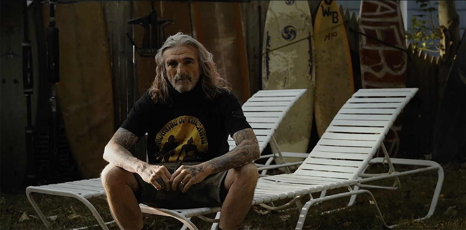 Photo of a man with long gray hair sitting on a white plastic reclining lawn chair with his elbows resting on his news. He wears shorts and a black and gold t-shirt and looks honest and content.