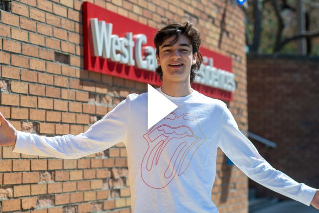 A photo of Will Loggia with his arms outstretched standing in front of a brick wall with a red sign that reads "West Campus Residences." A video play button is overlayed.