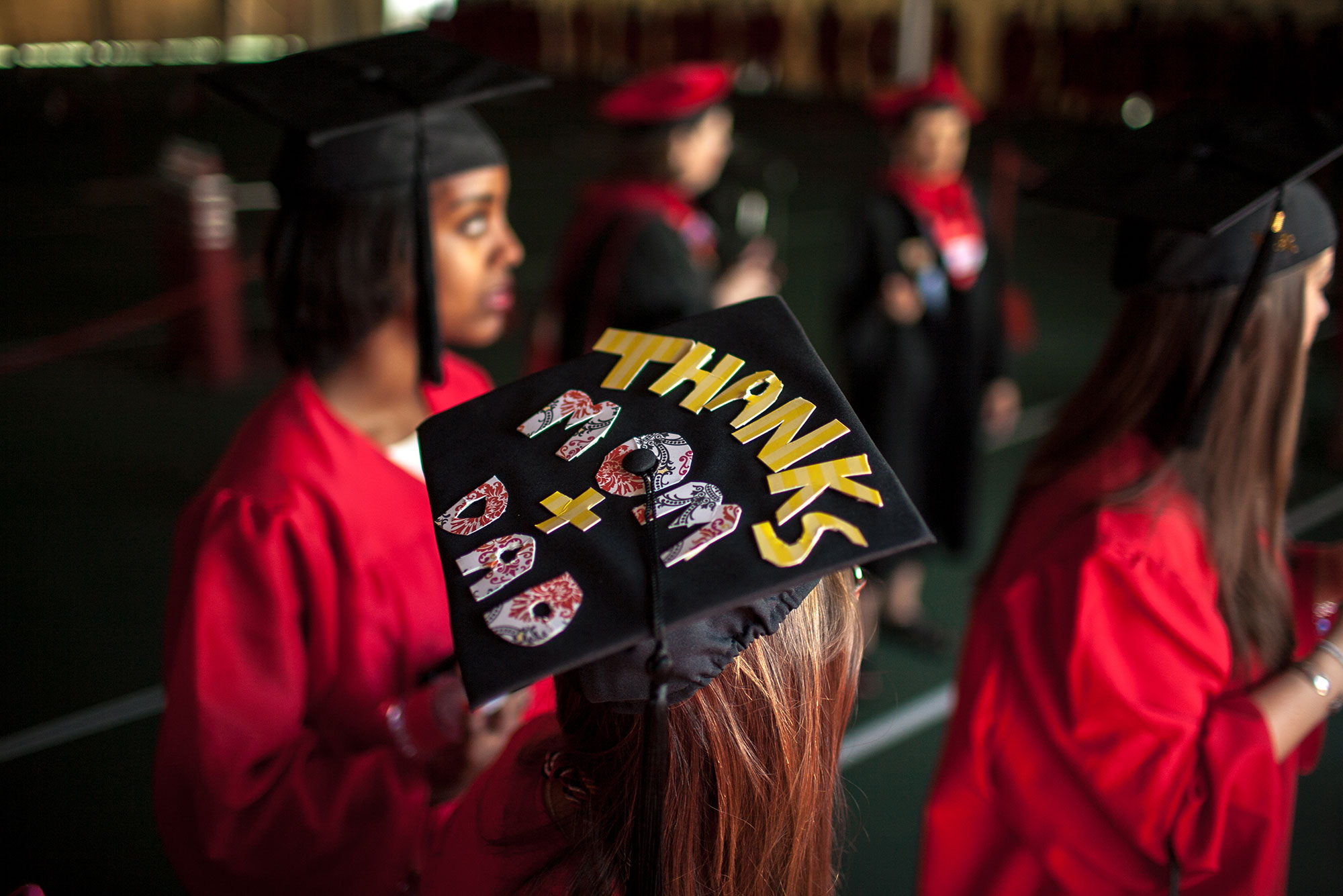 Photo of a group of students in cap and gowns, one has a mortar board that reads “thanks mom + dad” in cut out letters.