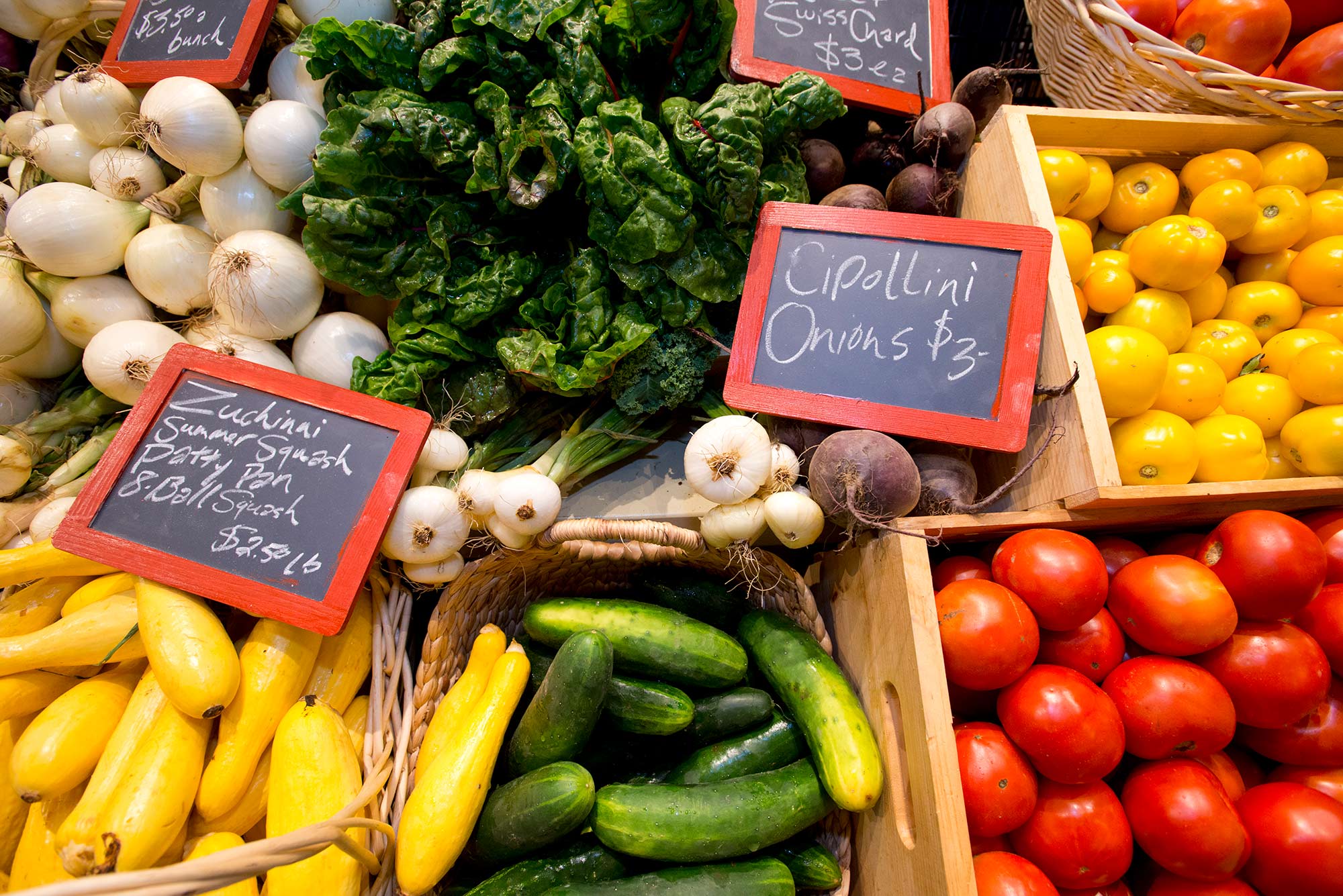 Photo of baskets and wooden boxes of fresh produce, taken at a slight angle from above. The photo includes white onion, cipolini onions, yellow tomatoes, yellow squash, red heirloom tomatoes and cucumbers. A few chalkboard signs are scattered on top of the produce and read “Zucchini, summer squash, patty pan, 8 ball squash $2.50/lb” and “cipollini onions. $3, swiss chard $3/lb.”