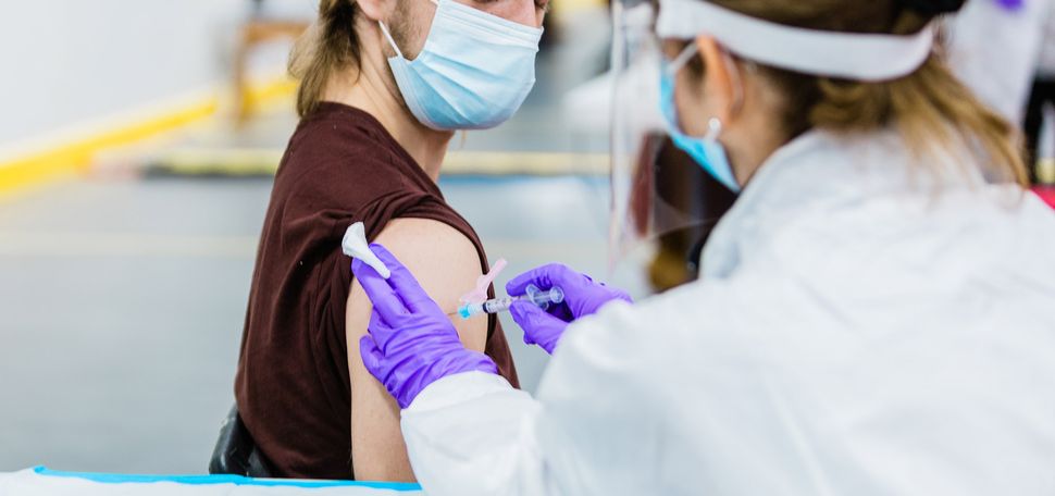 Photo of Ian Forcey, in a brown beanie, looking down at his shoulder, as a medical provider in a face shield, mask and purple gloves gives him a vaccine.