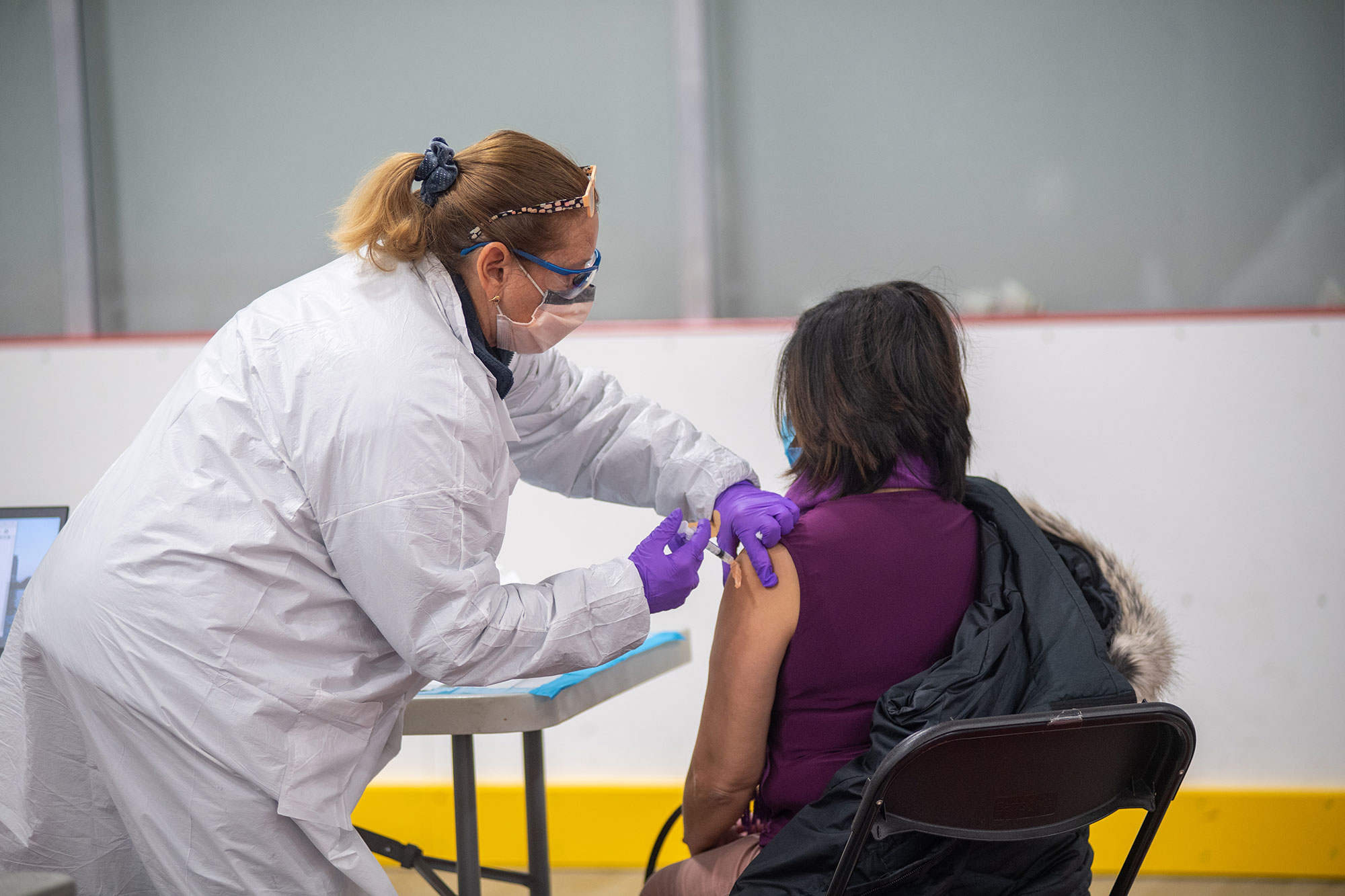 Photo of Terry Del Prato, RMA, in a white lab coat and purple glove, giving Yolanda Barros, seen from behind, with her jacket off one of her shoulders, receiving the Moderna COVID-19 vaccine January 20 at the vaccine clinic at FitRec.