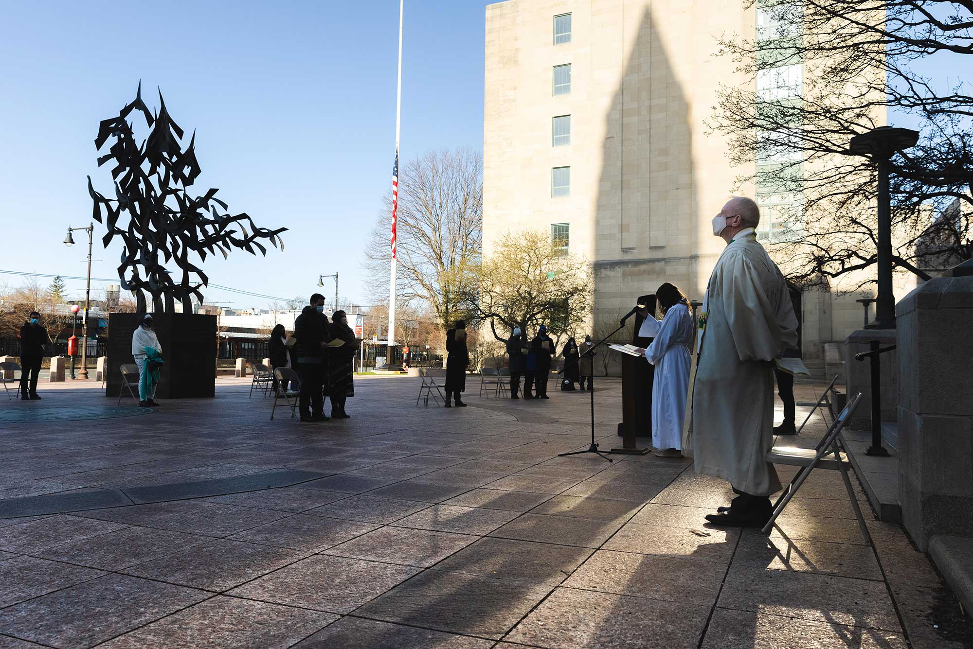 Photo of Rev. Robert Hill, dean of Marsh Chapel, standing with his hands behind his back looking out towards Marsh Plaza where a few groups sit in separated pods of folding chairs. Marsh Chapel’s interdenominational Easter Sunday sunrise vigil took place outdoors on Marsh Plaza April 4; the sky is light blue.