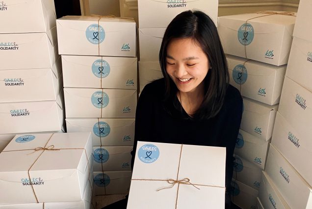 Photo of Sonia Su (COM’15), founder and executive director of Kits to Heart, surrounded by stacks of with kits, white boxes with twine bows, ready to be delivered. She holds one in her arms out in front of her and smiles and looks down. She has shoulder length black hair and wears a black sweater.