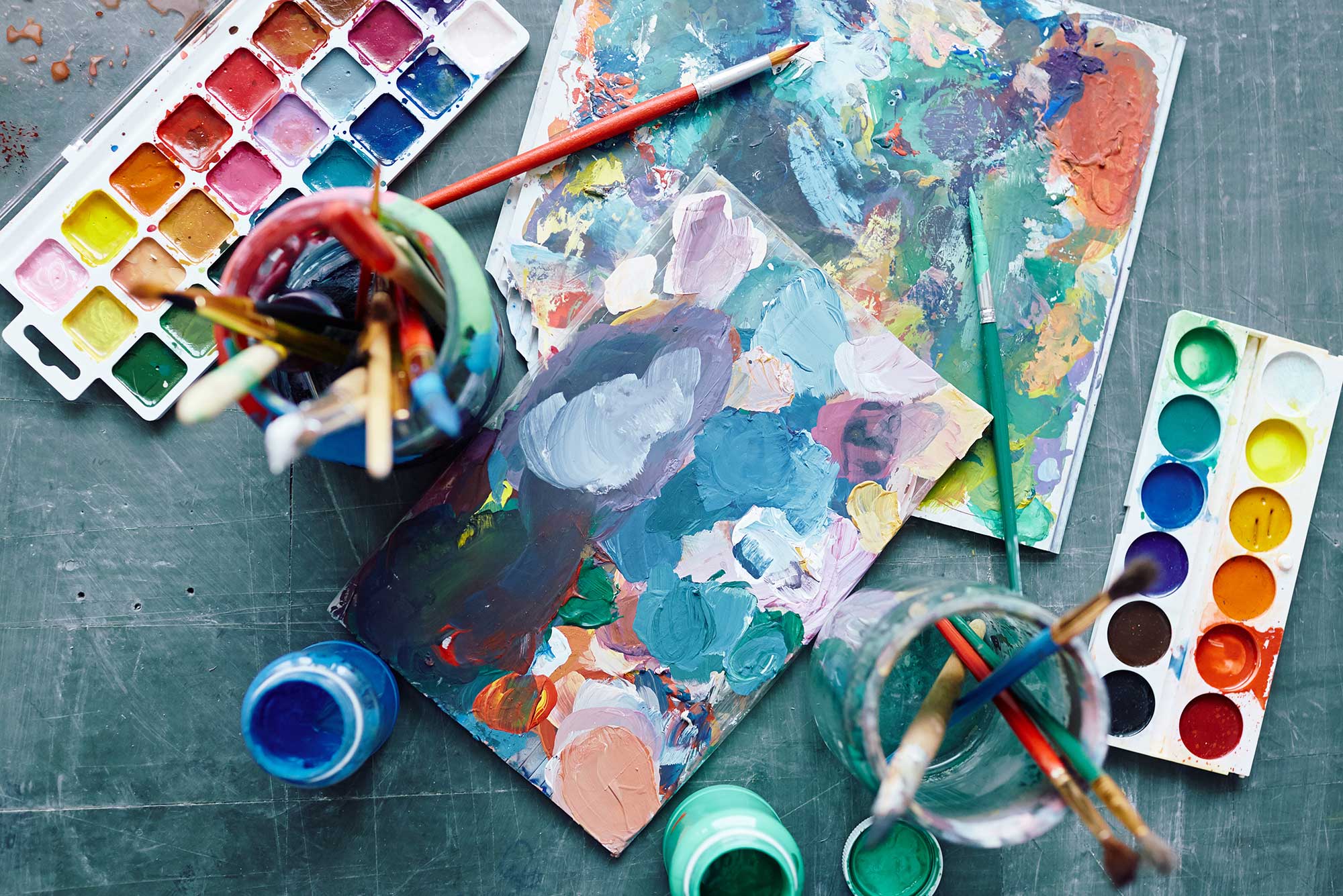 Photo of paint supplies, including brushes in water, color palettes, and a canvas with paint blotches and paint bottles resting on a desk.